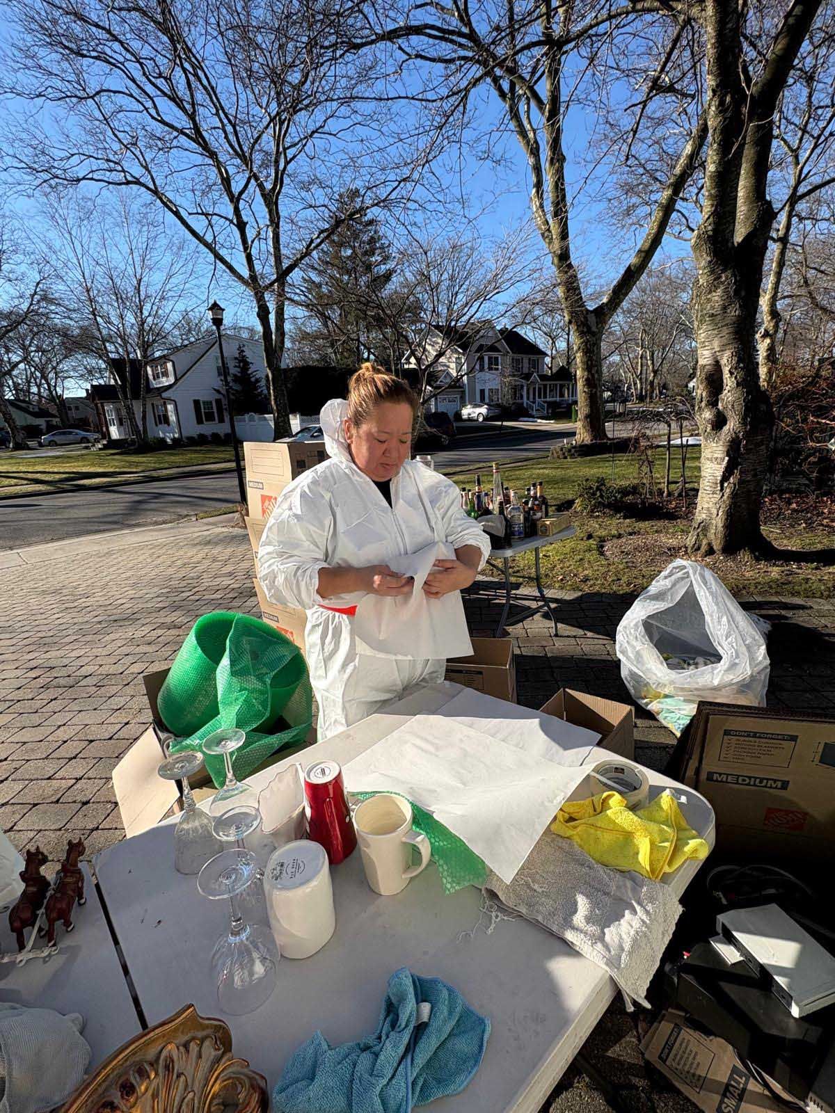 A person wearing a white protective suit stands at a folding table, wrapping items in paper outdoors on a sunny day.