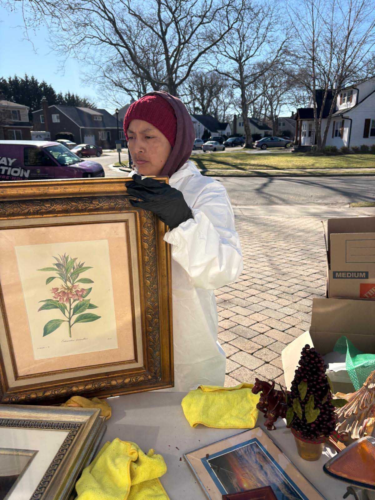 A person in a protective suit and black gloves holds a framed botanical print outside on a sunny day near a table.