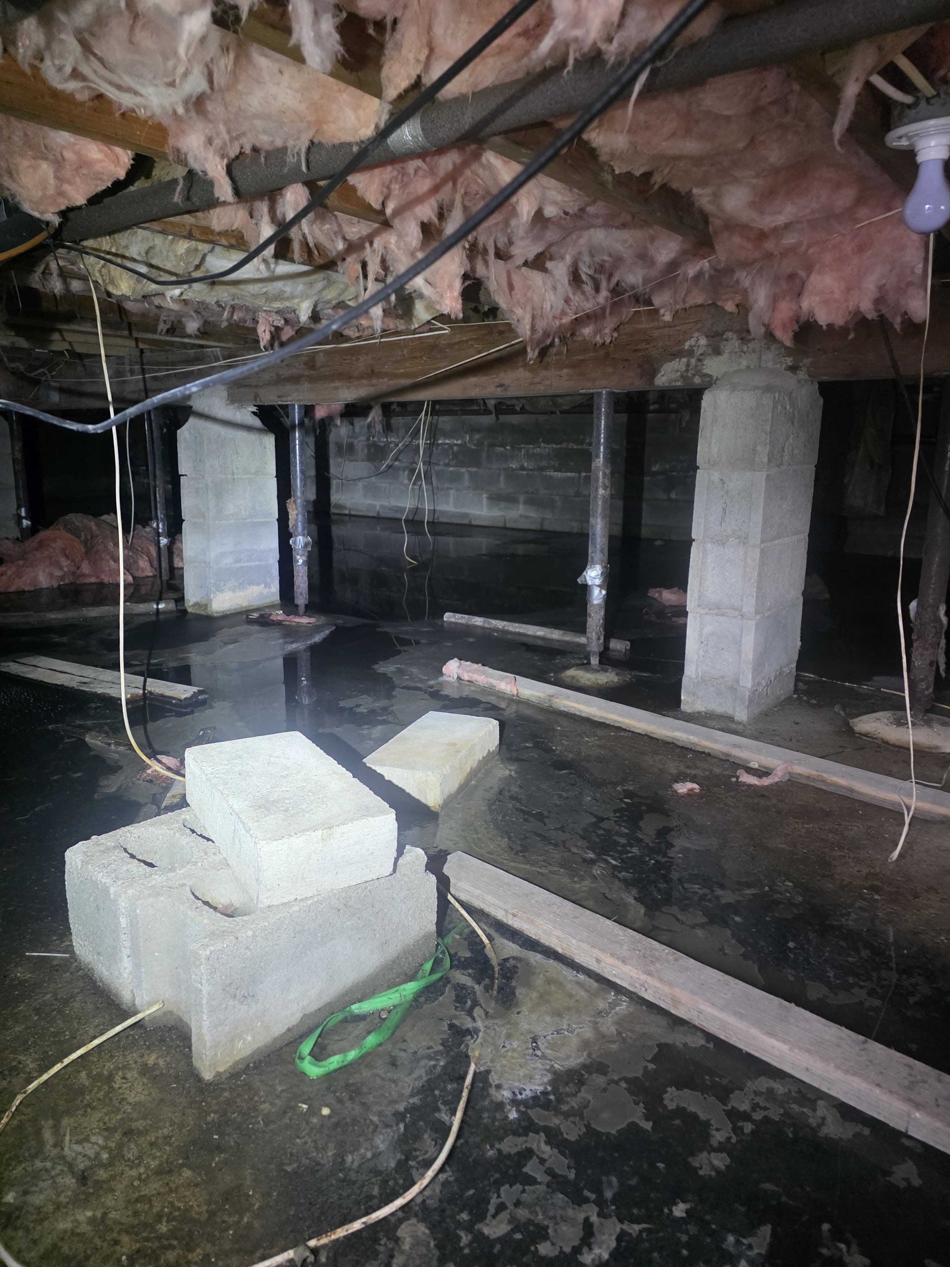 A dark, damp crawl space with concrete piers, exposed wooden floor joists, hanging insulation, and standing water.