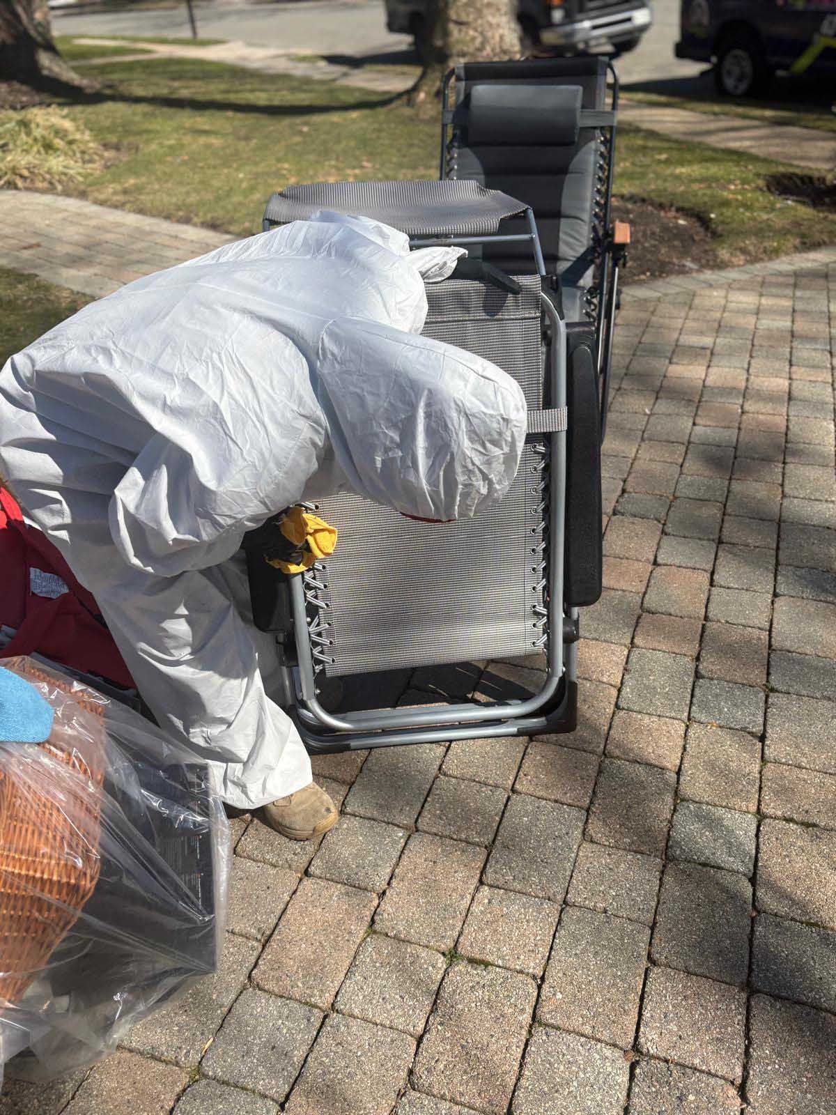 A person in a white protective suit inspects a gray folding lounge chair on a paved driveway.