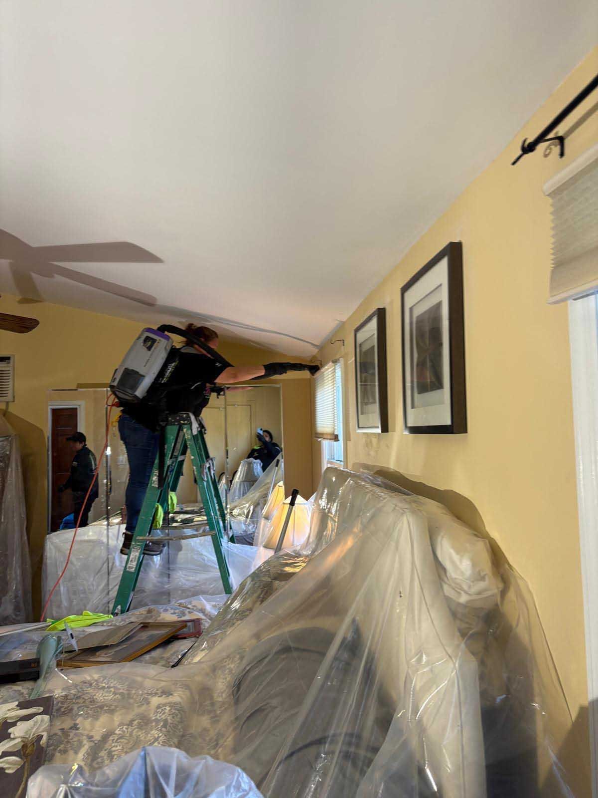 A worker on a ladder uses a paint sprayer on a wall in a yellow room protected by plastic sheeting.