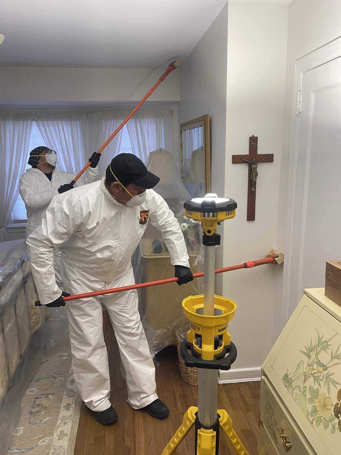 Two workers in white protective suits and masks using specialized tools to perform work on walls in a home.