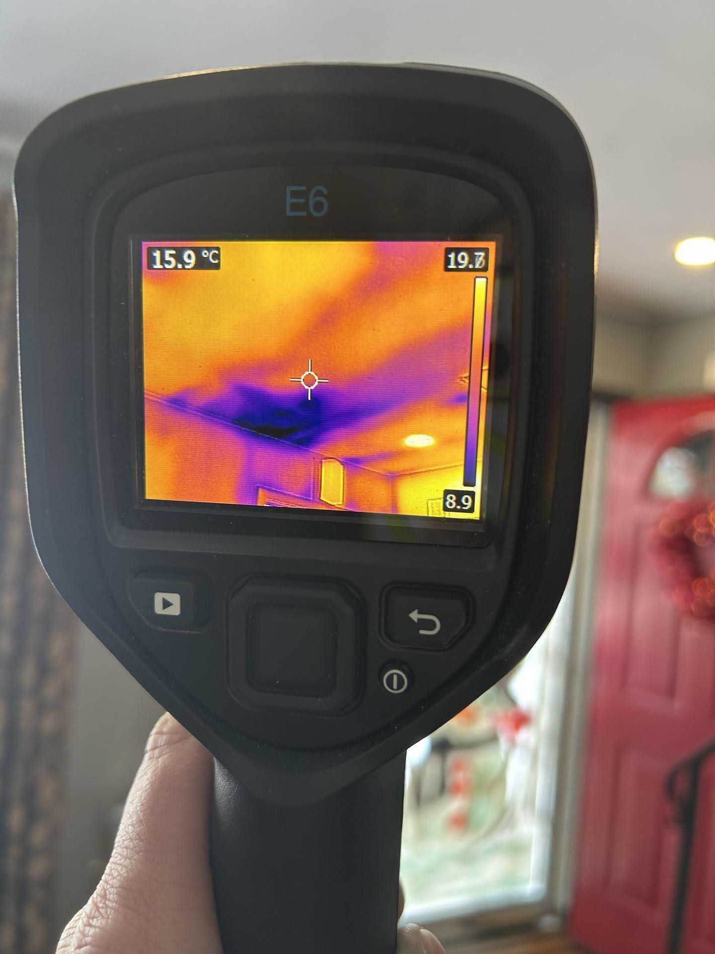 A thermal imaging camera held in a hand, displaying a ceiling with purple and orange heat patterns indicating insulation.