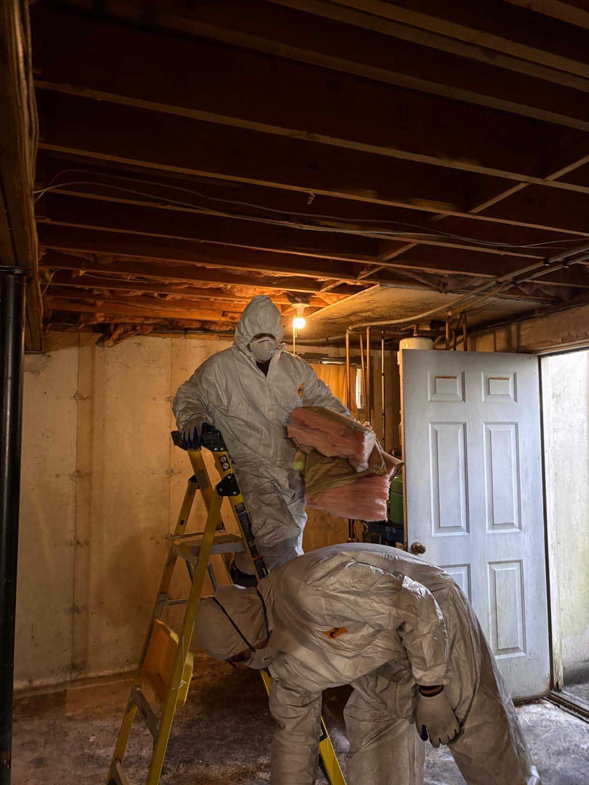 Two people in white protective hazmat suits working on insulation in an unfinished basement with exposed wooden joists.