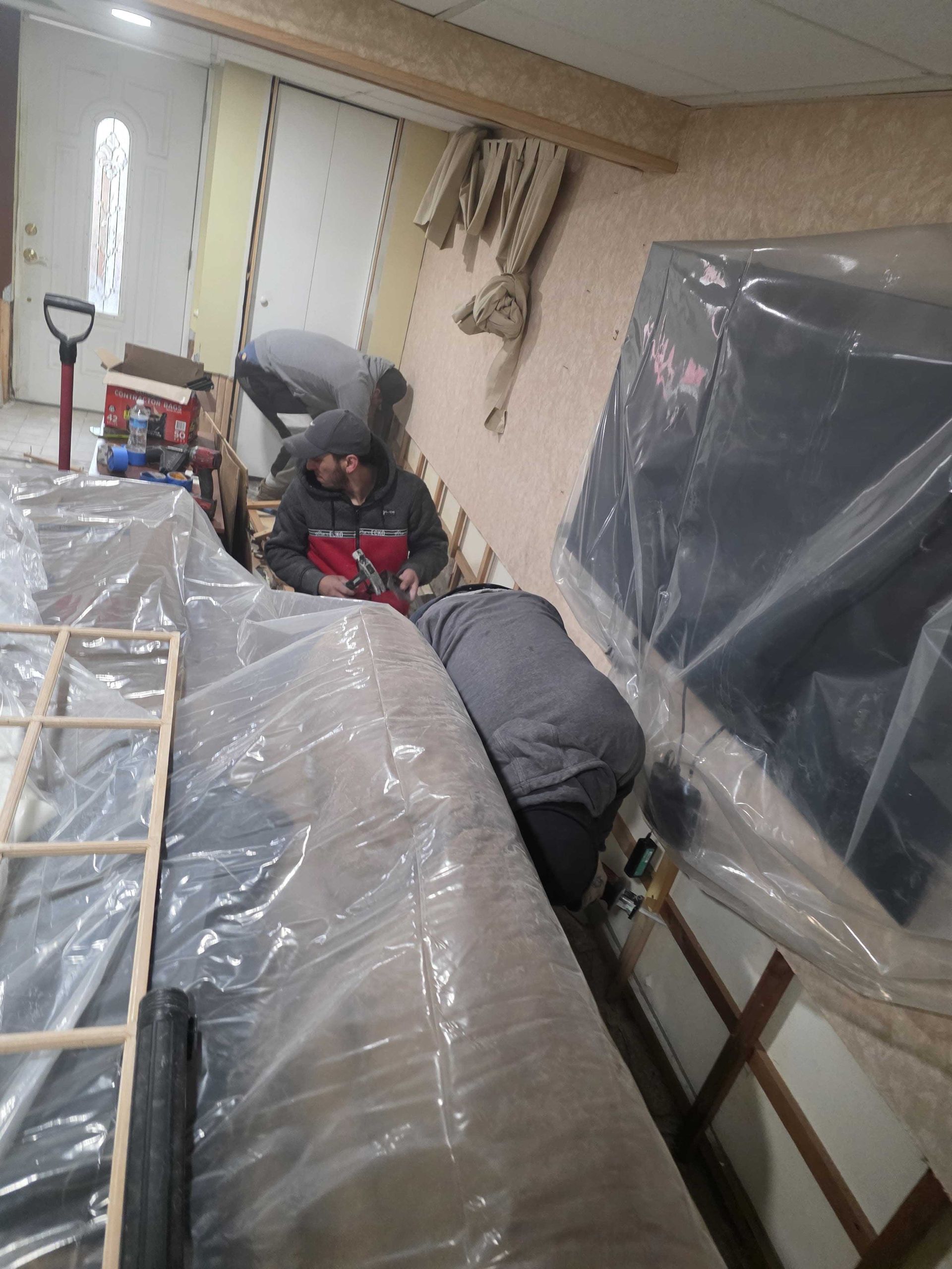 Two people work on a wall renovation in a room covered with plastic sheeting, using tools to attach wood framing.
