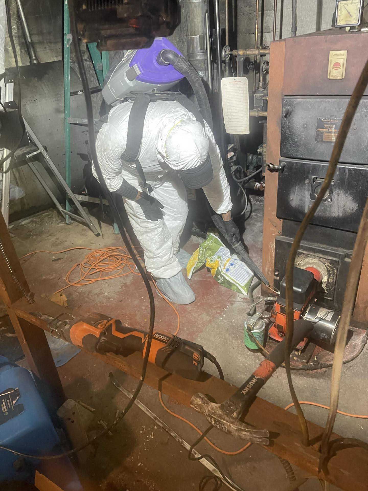 A person in protective coveralls and a backpack vacuum cleans debris near a boiler in a dimly lit utility room.