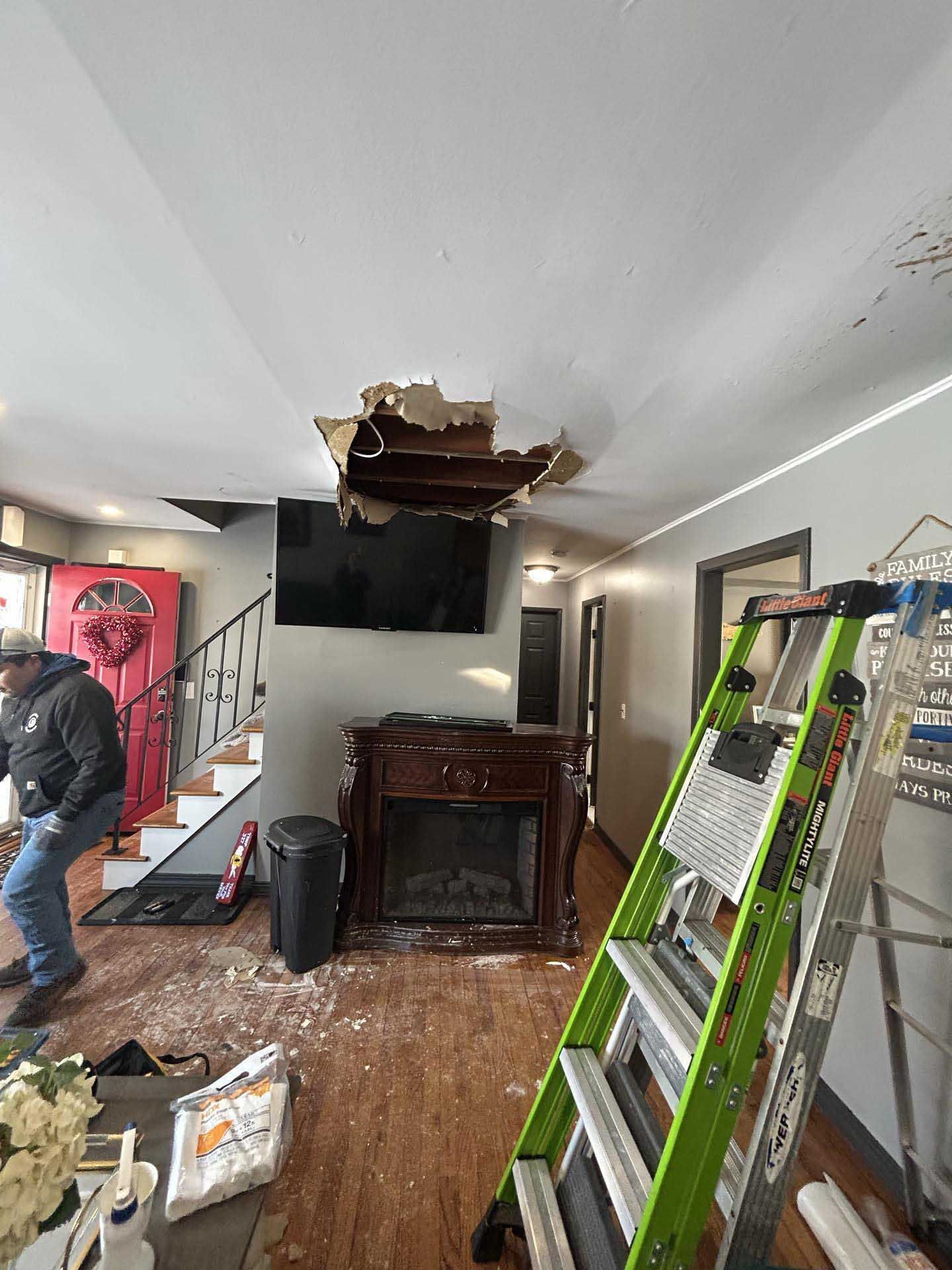 A worker stands near a fireplace in a room with a large, jagged hole in the ceiling above a wall-mounted TV.