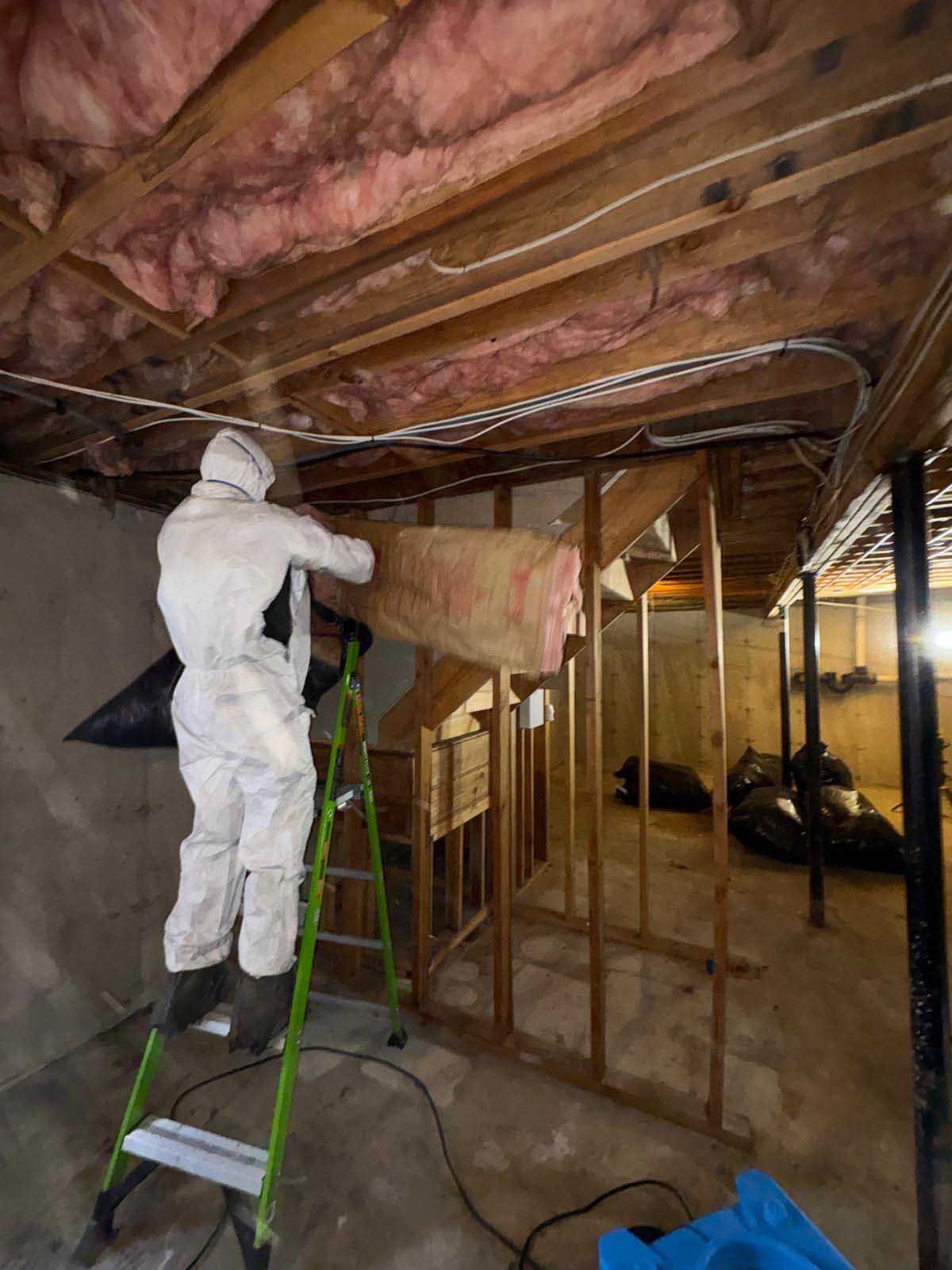 A person in protective coveralls stands on a ladder, installing pink fiberglass insulation between basement ceiling joists.