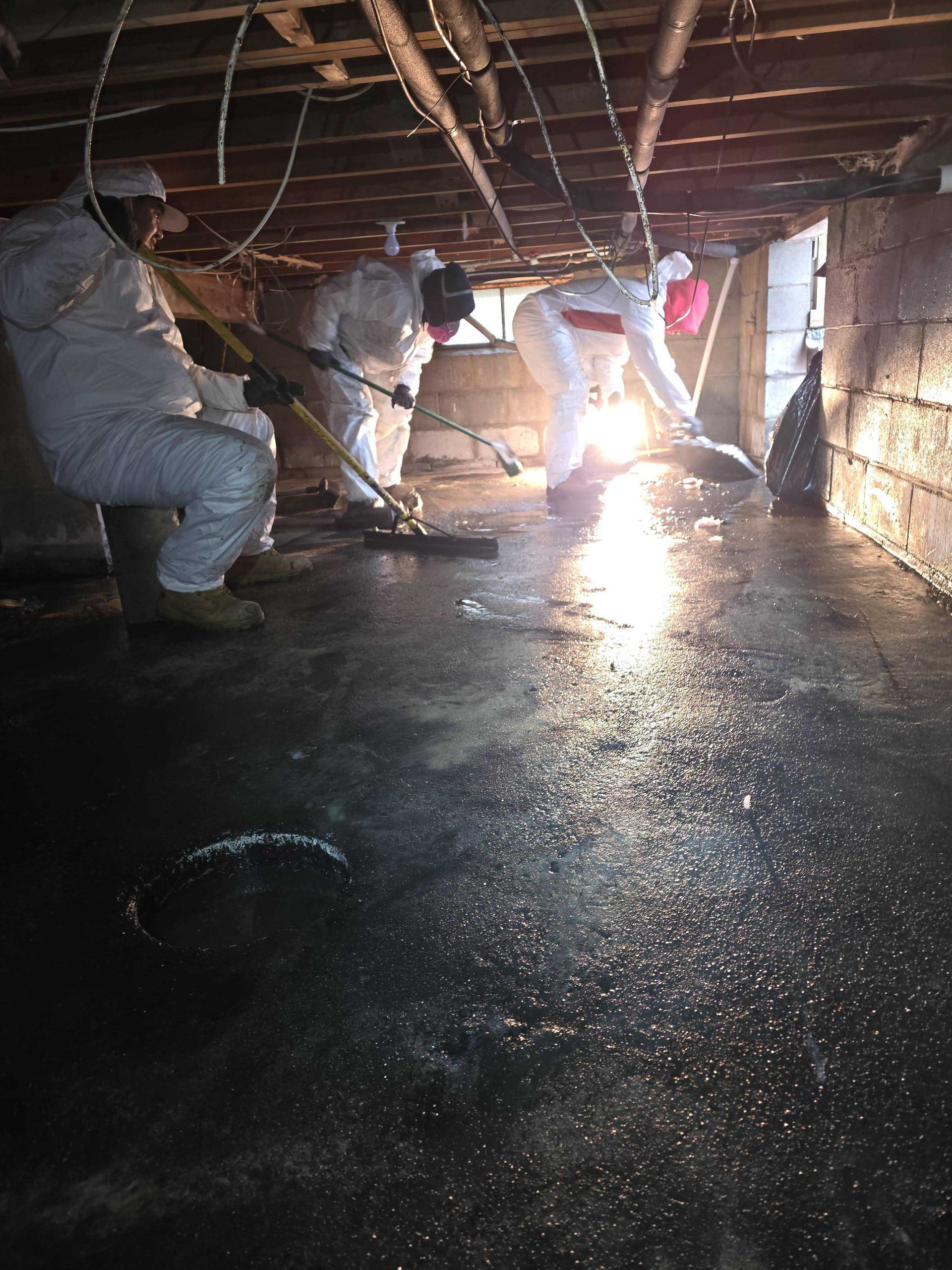 Workers in white protective suits clean a damp, low-ceilinged basement with reflective, dark, wet floors.