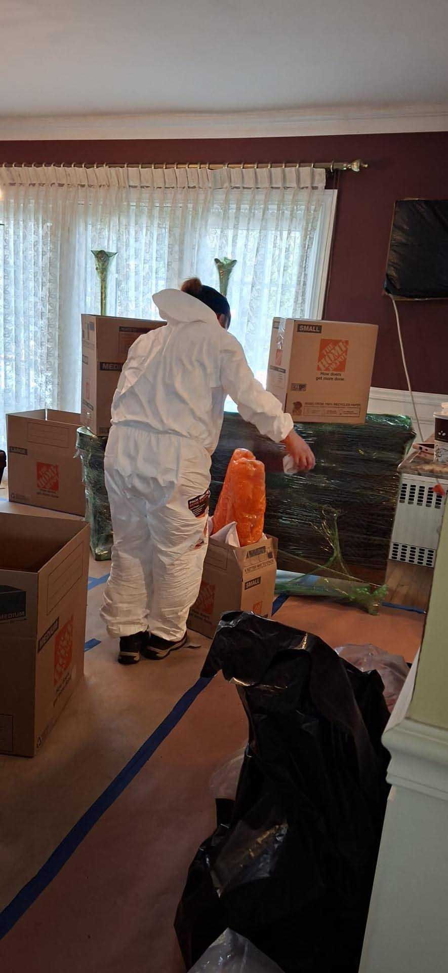 A person in white protective gear packing items into cardboard boxes inside a room filled with moving supplies.