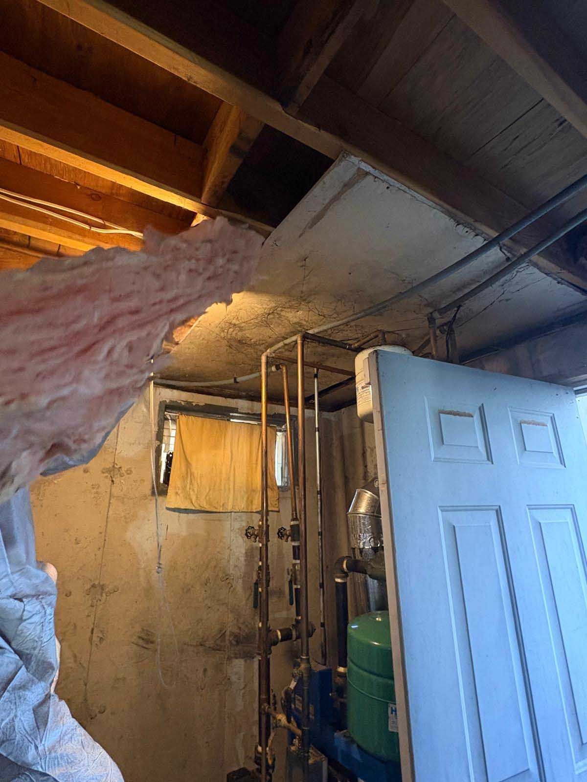 Basement corner showing exposed wooden ceiling joists, pink fiberglass insulation, copper pipes, and a door.
