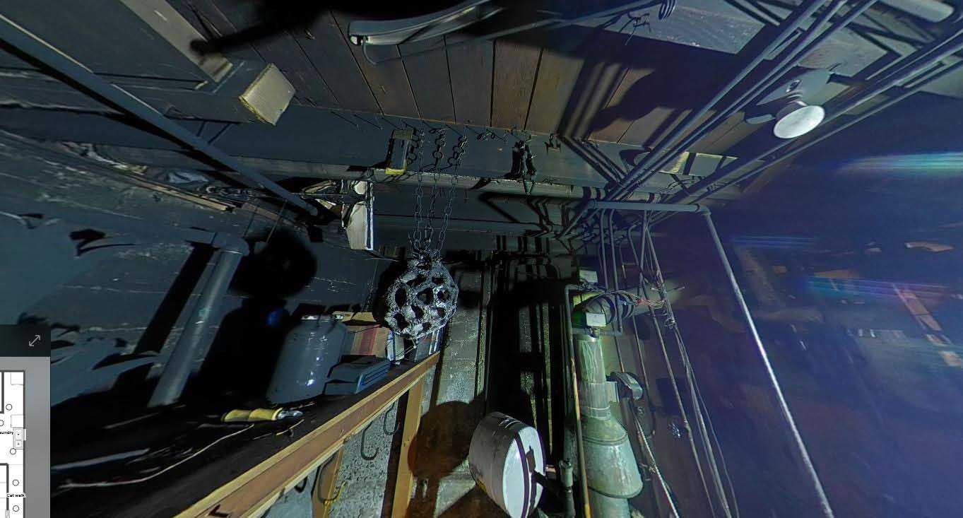 A dimly lit, cramped interior space of a historic ship, featuring exposed pipes, wiring, and metal structures.