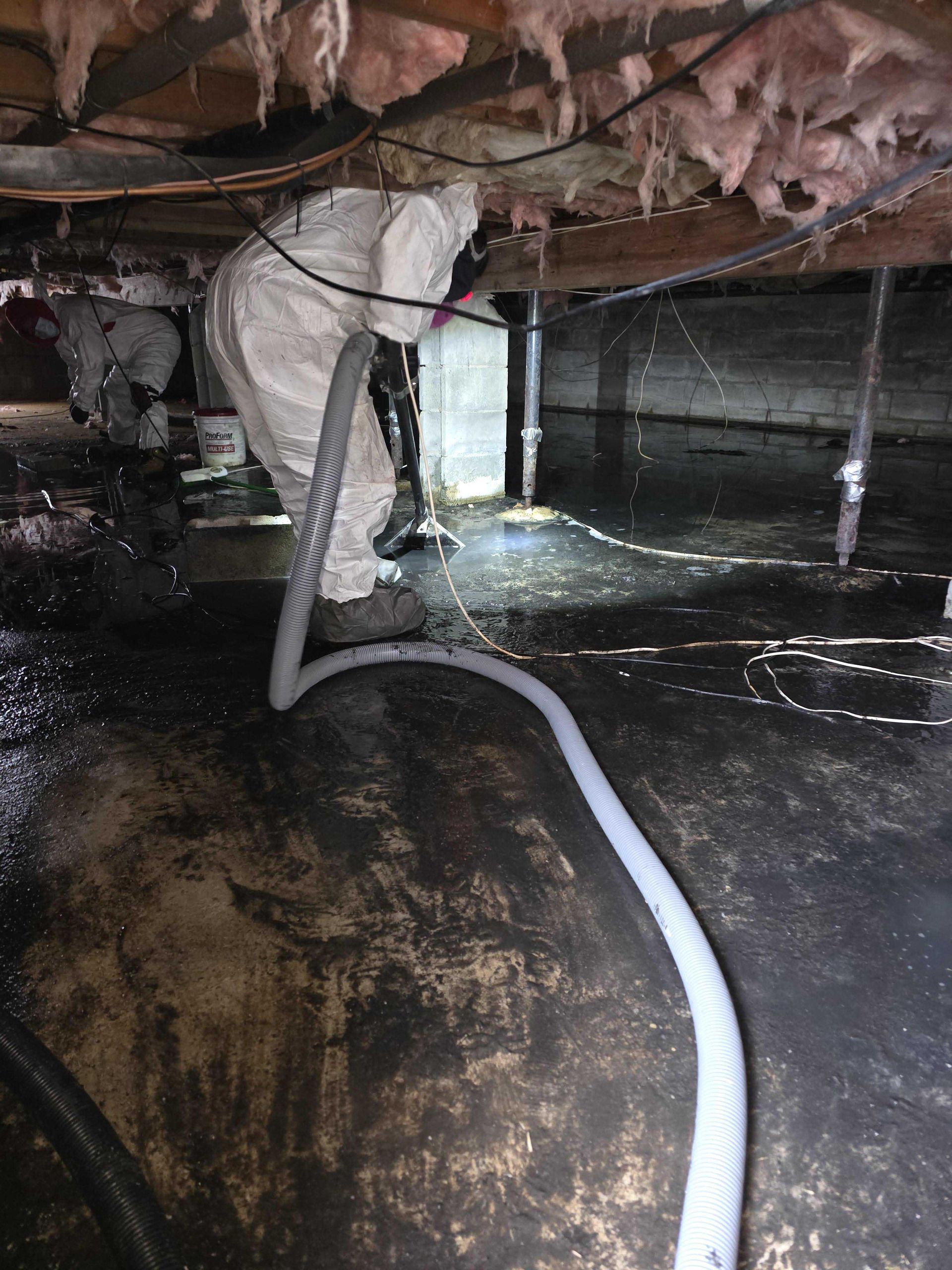 A worker in a white protective suit uses a long hose to clean a flooded crawl space under a house with exposed insulation.