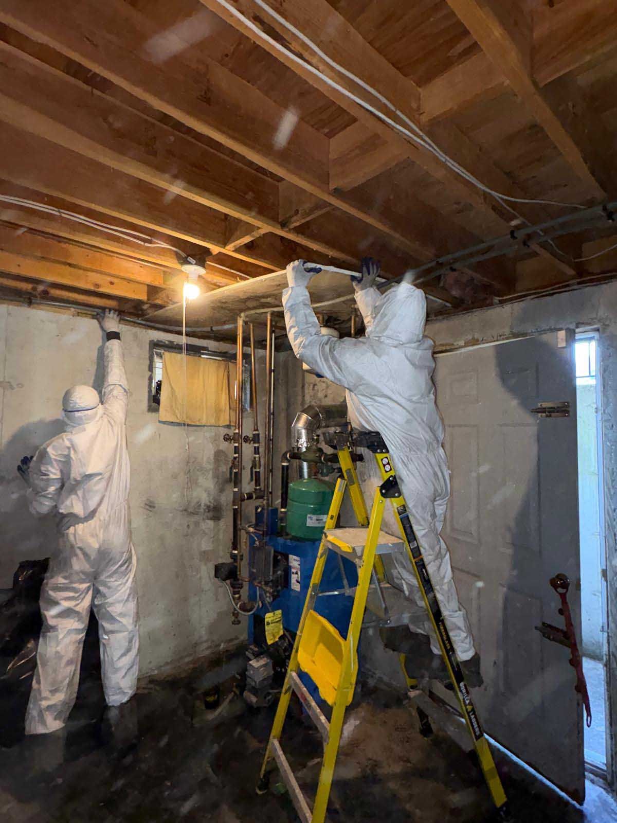 Two workers in white hazmat suits perform remediation work on the ceiling of a basement utility room.