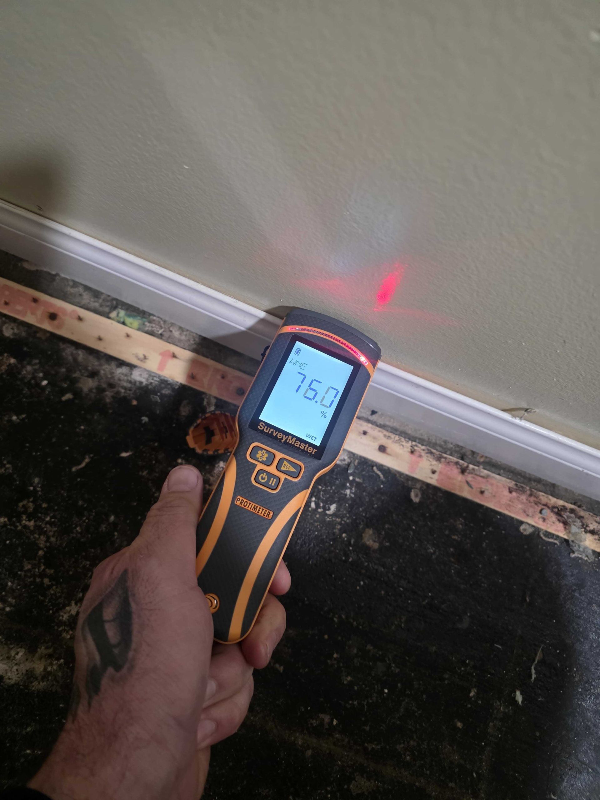 A hand holds an infrared thermometer displaying 75.0 degrees, pointed at a wall near a floor with dark flooring.