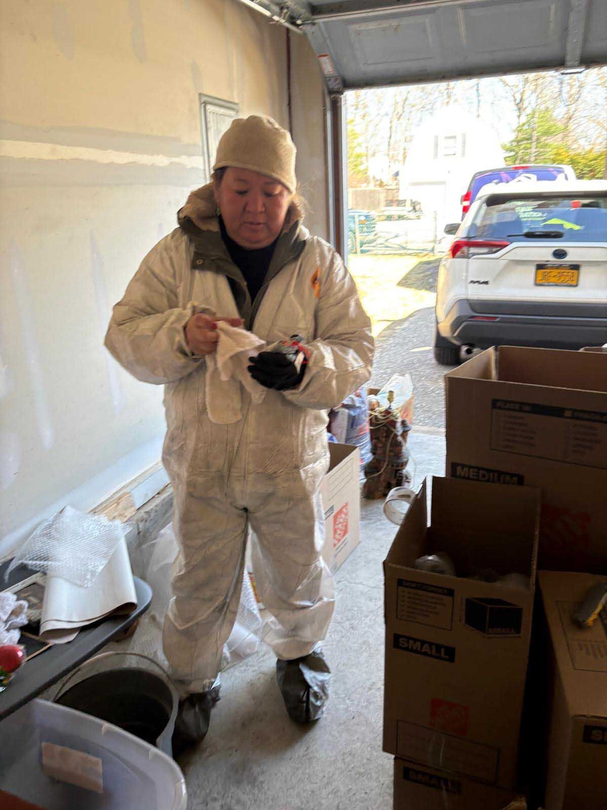 A person in protective white coveralls and a hat wipes an item while standing in a garage with cardboard packing boxes.