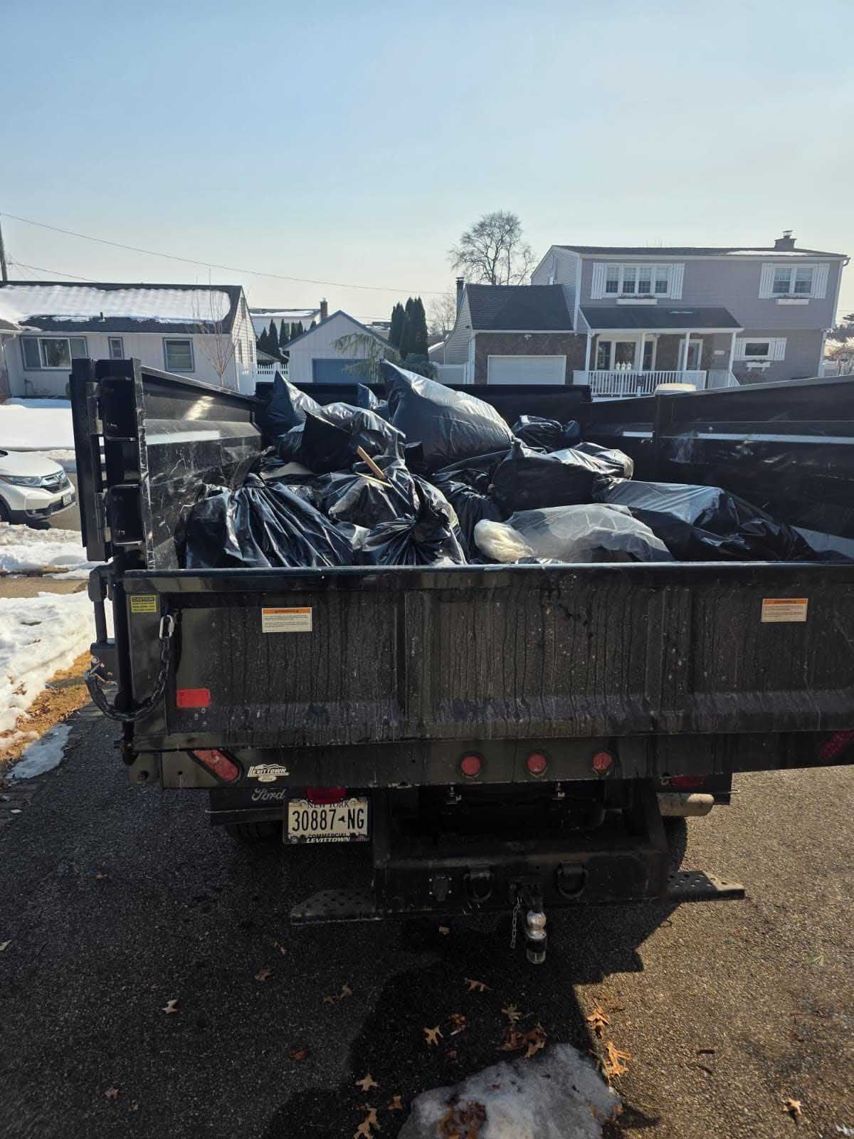 A black dump trailer filled with trash bags parked on a street near houses on a sunny, winter day.