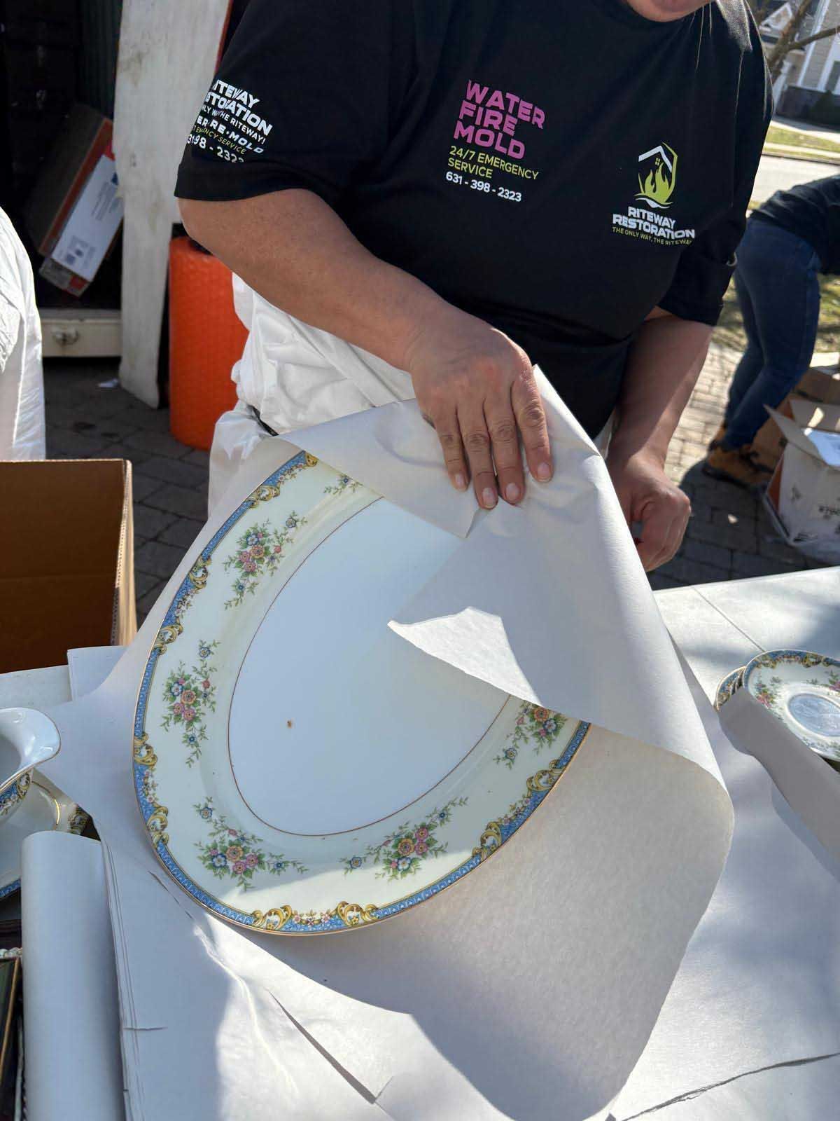 A person wearing a black uniform wraps a decorative, floral-patterned ceramic dish in white paper outdoors.