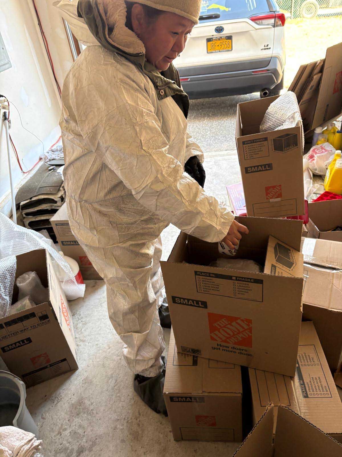 A person in a white protective suit reaches into a cardboard box in a garage near a parked white vehicle.