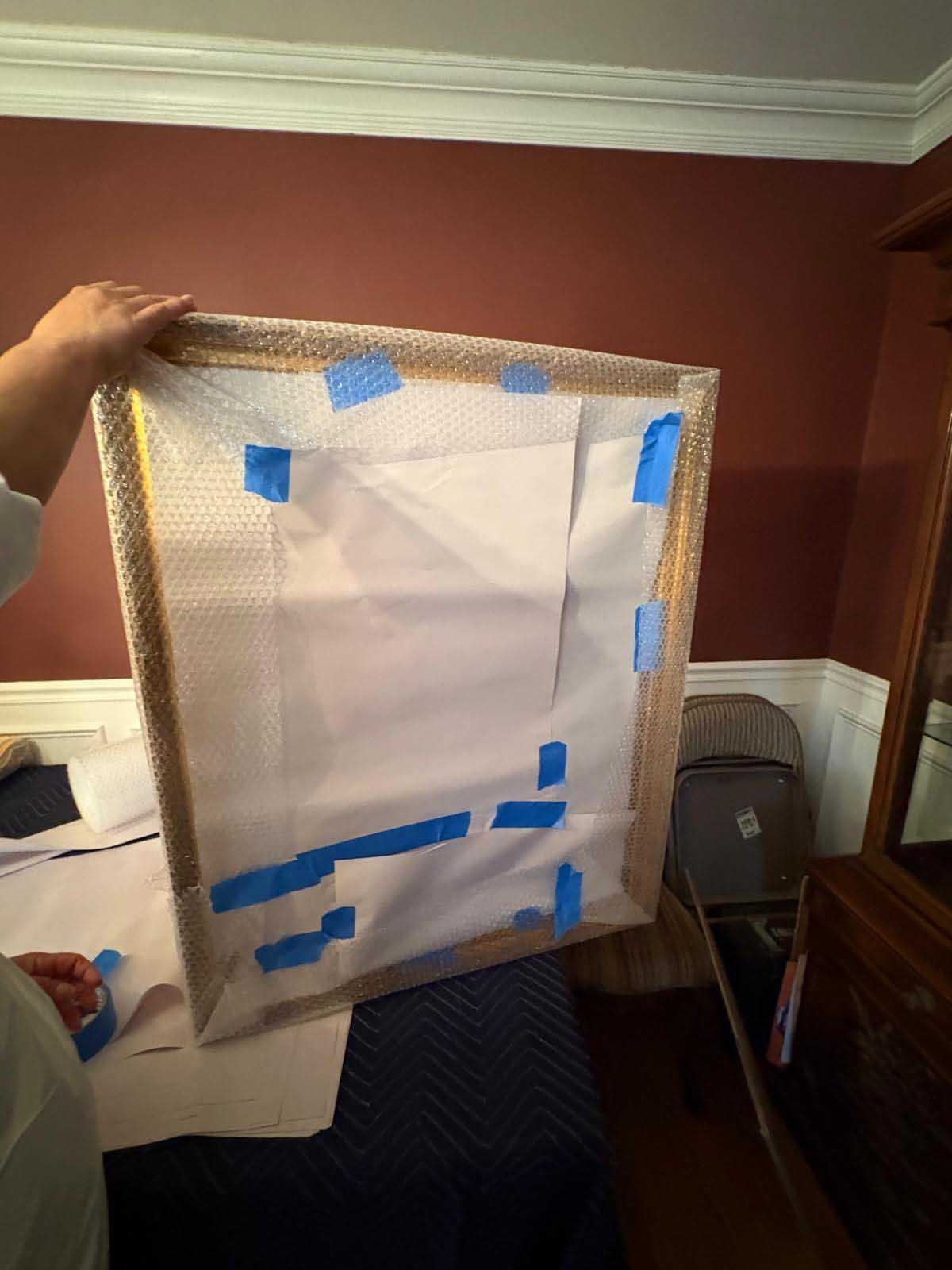 A person holds a large framed item wrapped in white paper and bubble wrap, secured with blue painter's tape.