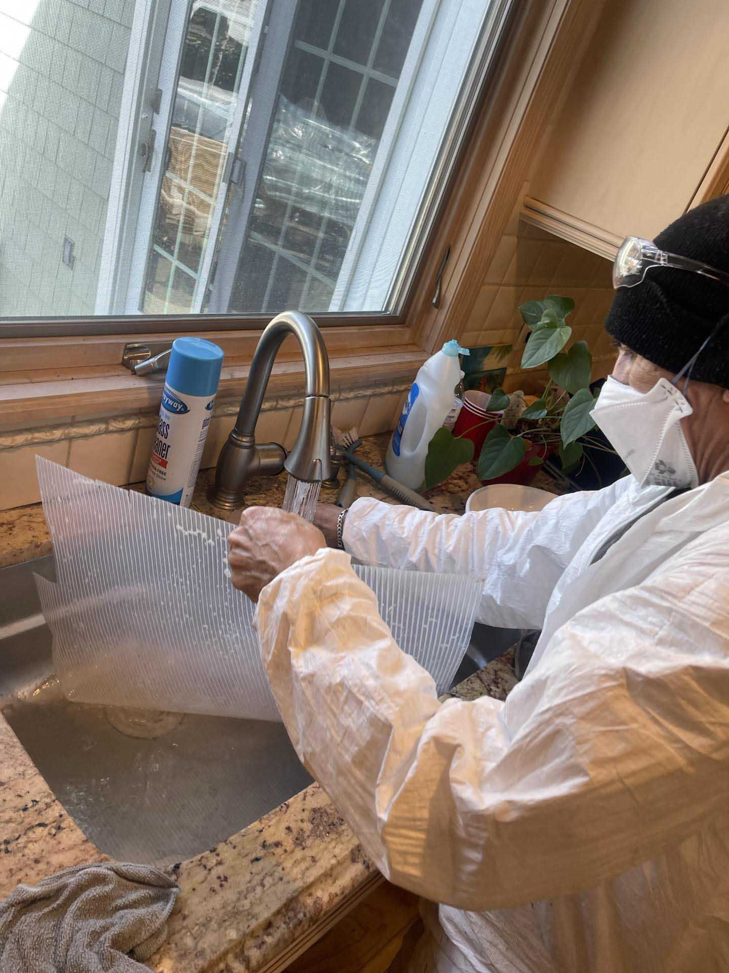 A person in a protective suit, mask, and beanie cleans a white patterned panel under a kitchen faucet.