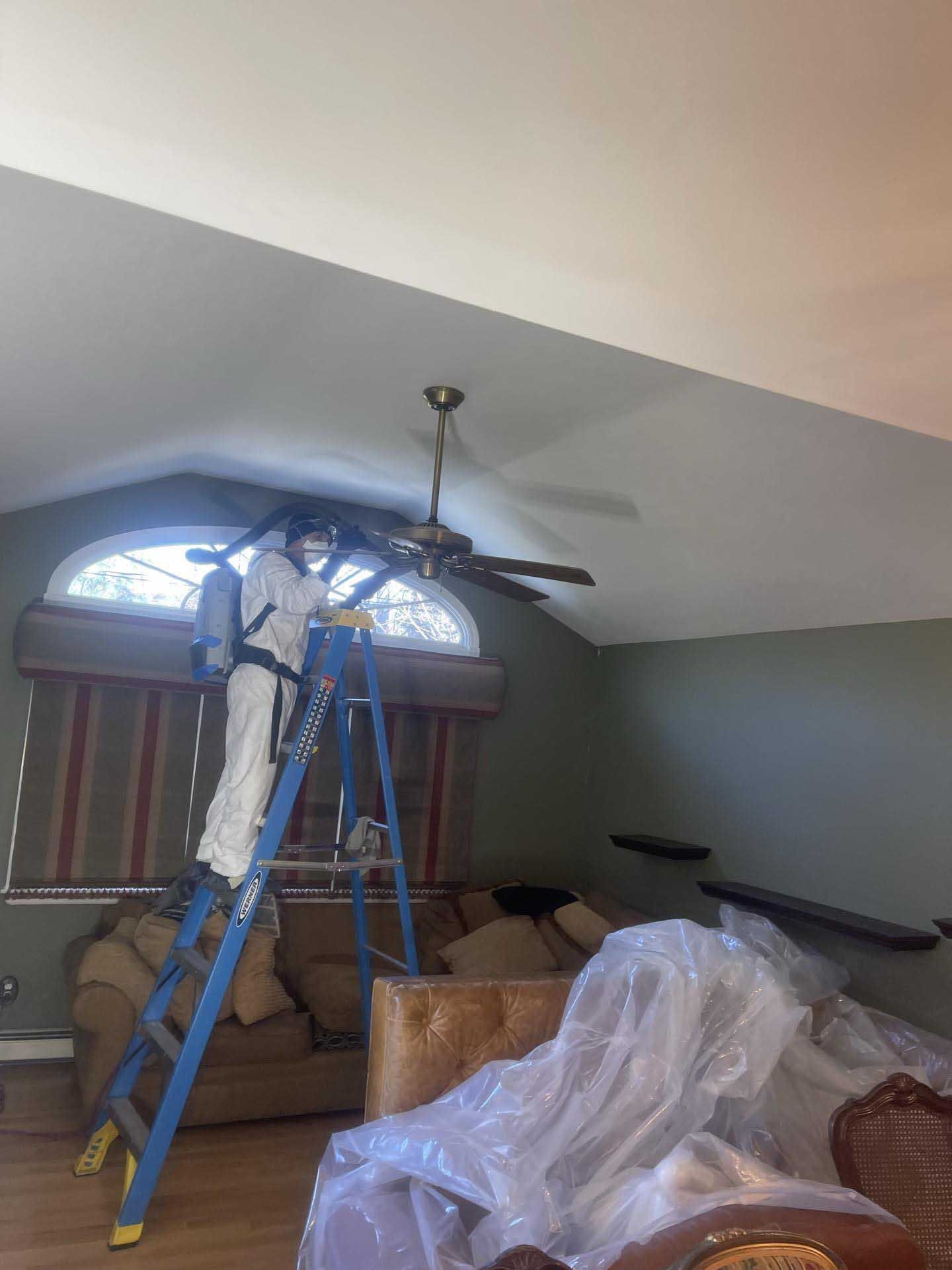 A worker in protective gear stands on a ladder cleaning a ceiling fan in a living room with covered furniture.