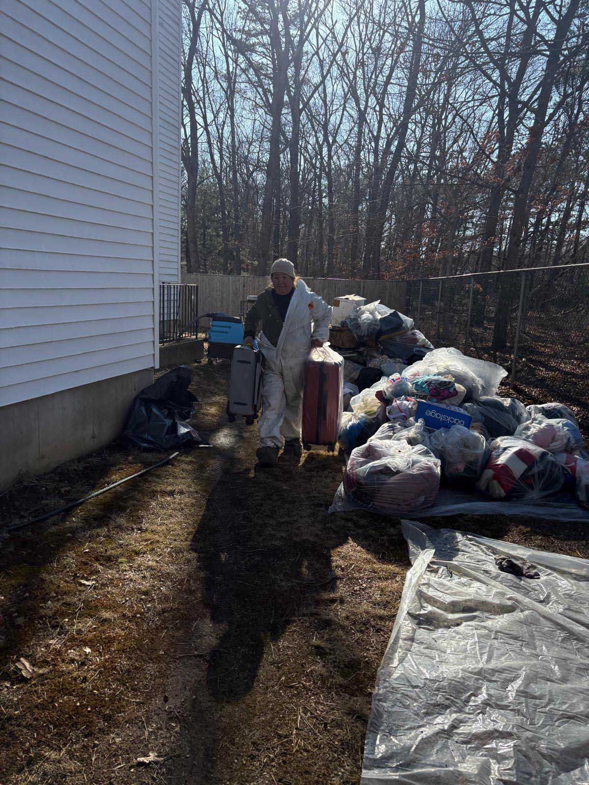 A person in a white hazmat suit moves two suitcases past a large pile of bags and debris outside a house.