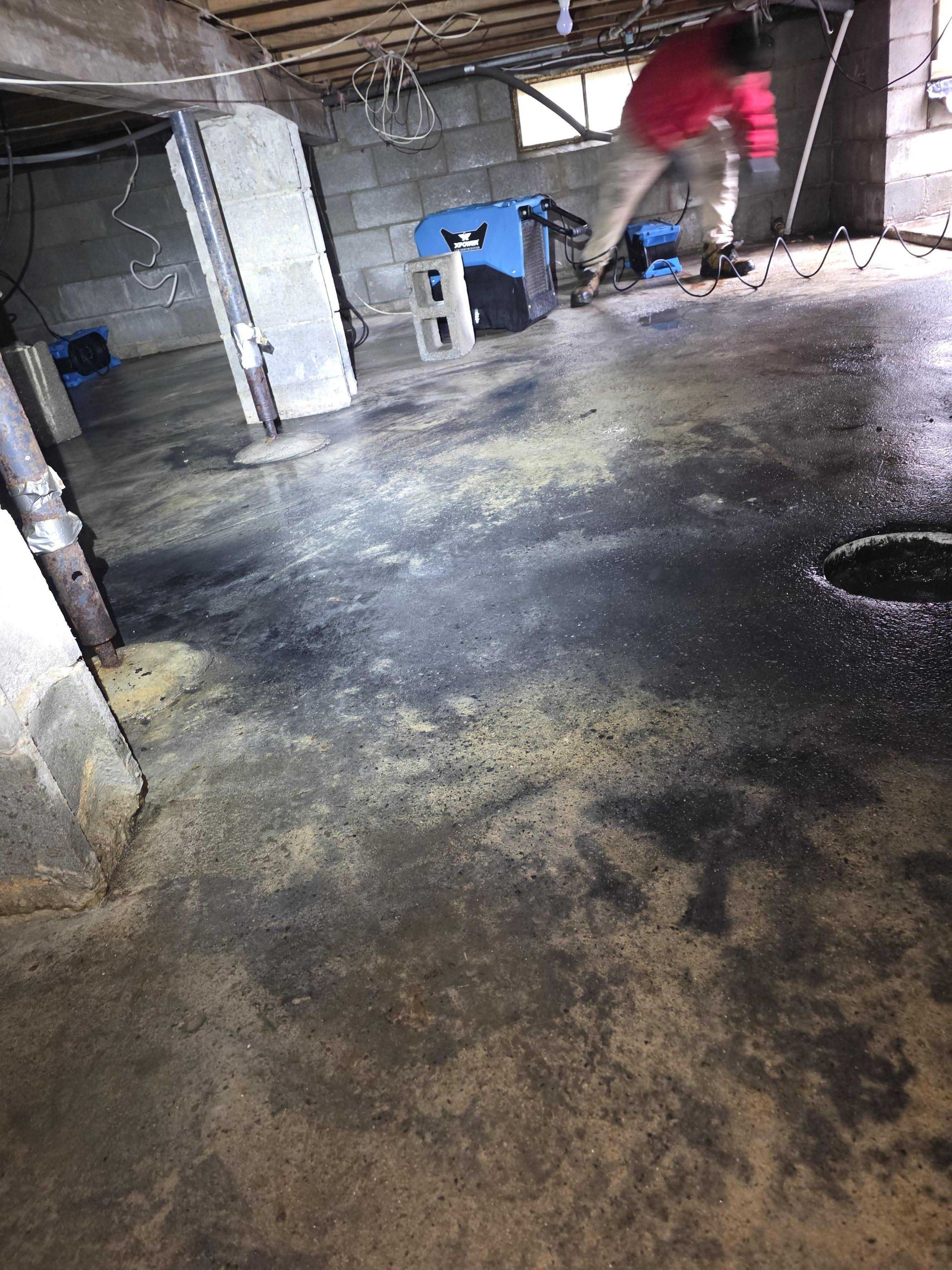 A person in a red jacket works in a dimly lit, damp concrete basement with industrial drying equipment.