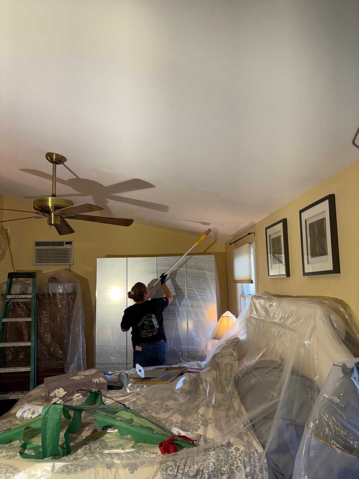 A person using a long-handled roller to paint a ceiling in a room with yellow walls and furniture covered in plastic.