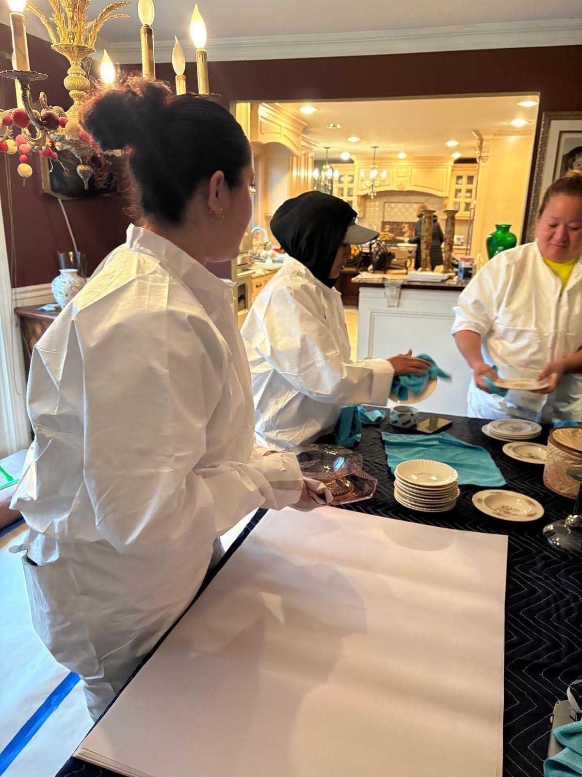 Three people in white protective coveralls stand around a table covered with a large white sheet, sorting items.