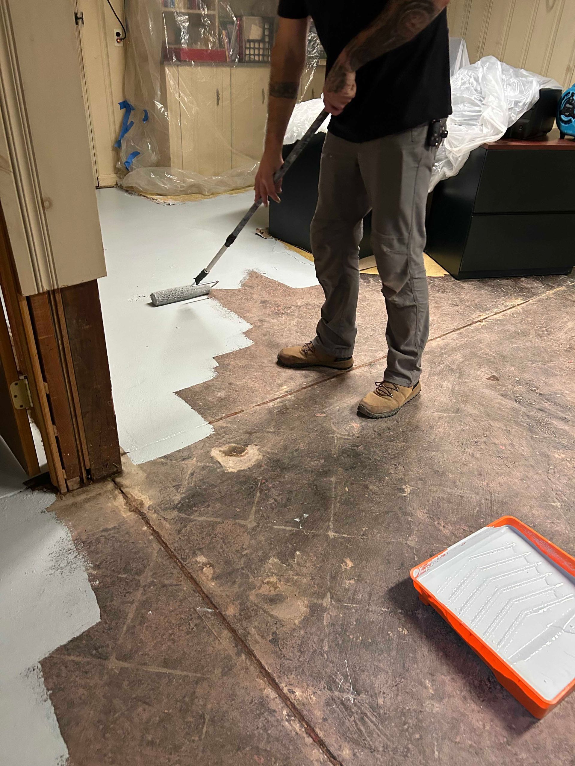 A person using a roller to apply light gray paint to a concrete floor in an indoor space with a paint tray nearby.