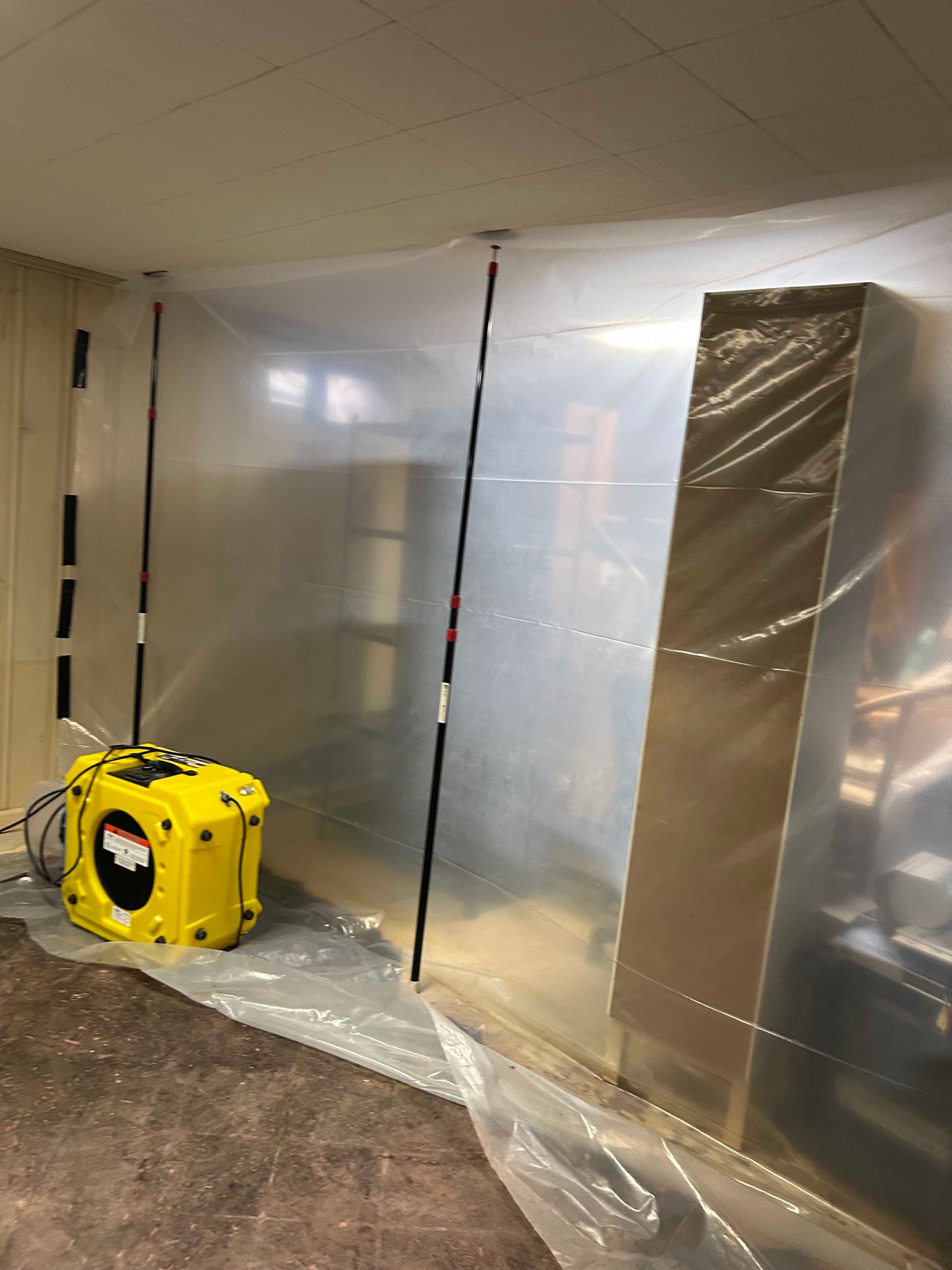 A yellow air scrubber sits on a plastic floor sheet next to a ceiling-high plastic wall barrier held up by poles.