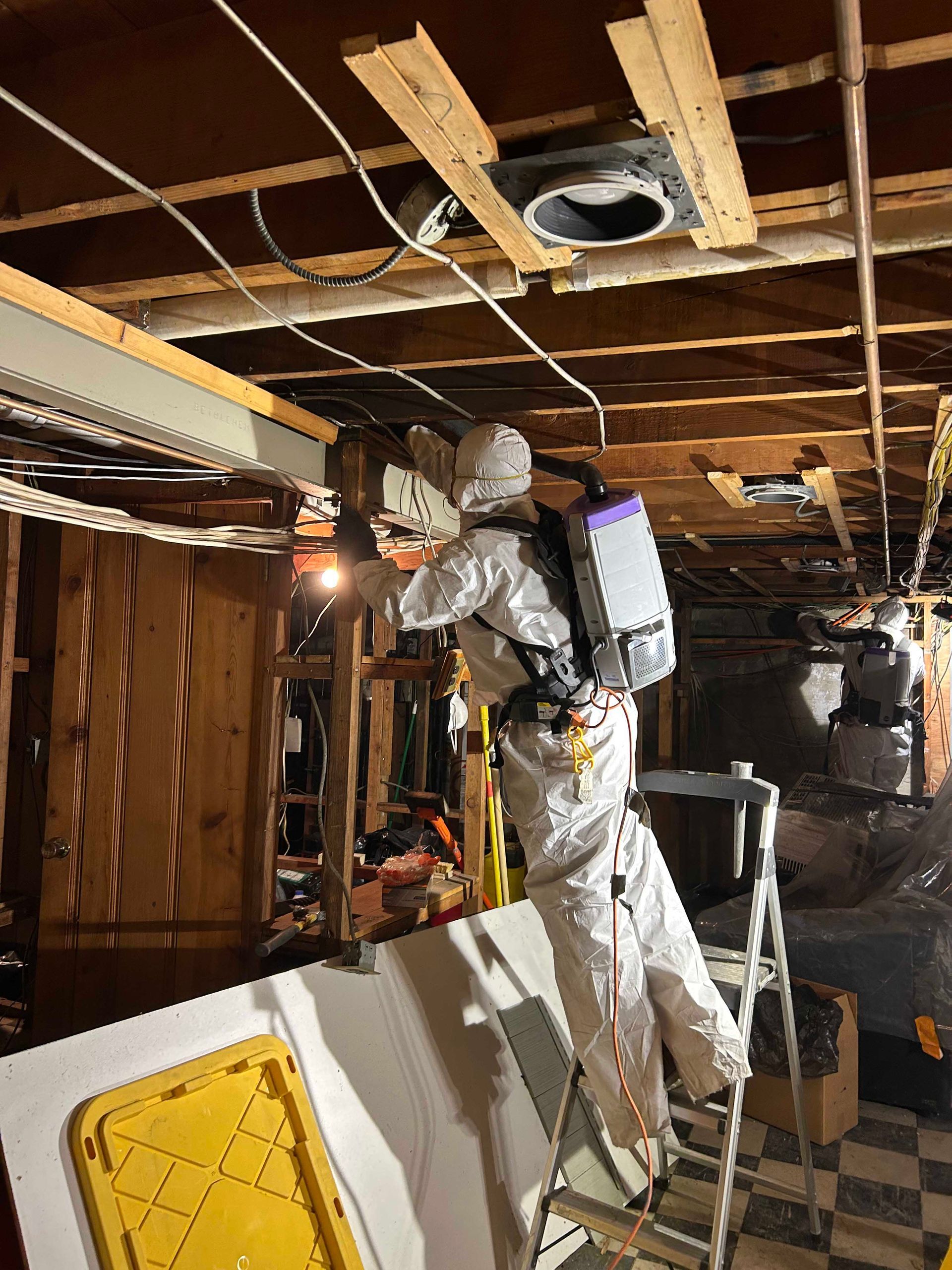 A person in white protective gear and a backpack vacuum cleans an unfinished ceiling in a room under renovation.