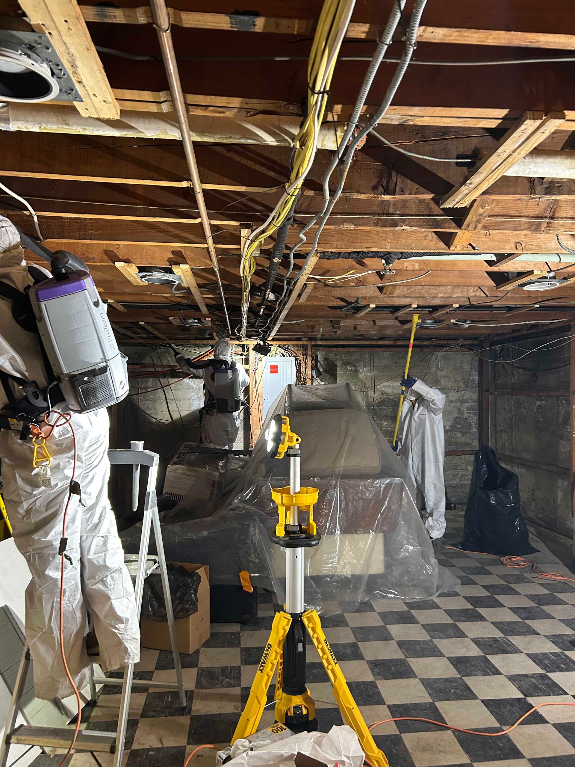 Two workers in protective suits remediate mold in a basement with exposed ceiling joists, equipment, and a tripod light.