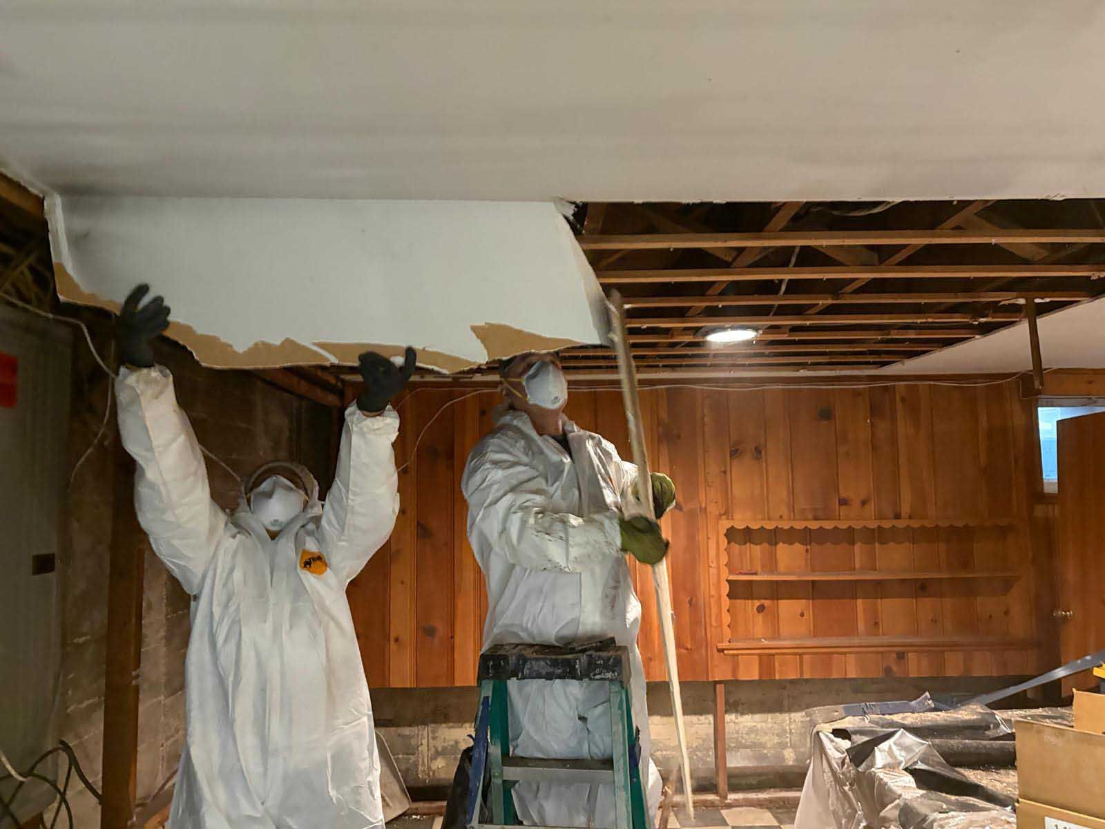 Two workers in protective white coveralls and masks remove ceiling panels in a rustic wood-paneled room.