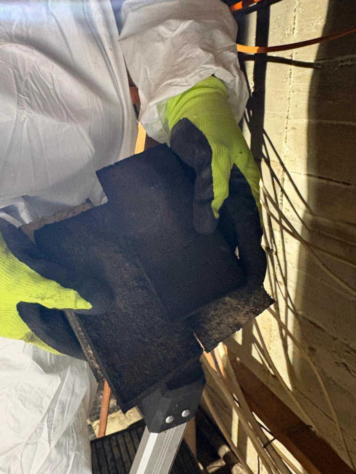 A person in a protective suit and gloves holds a piece of black insulation material inside a wooden building structure.