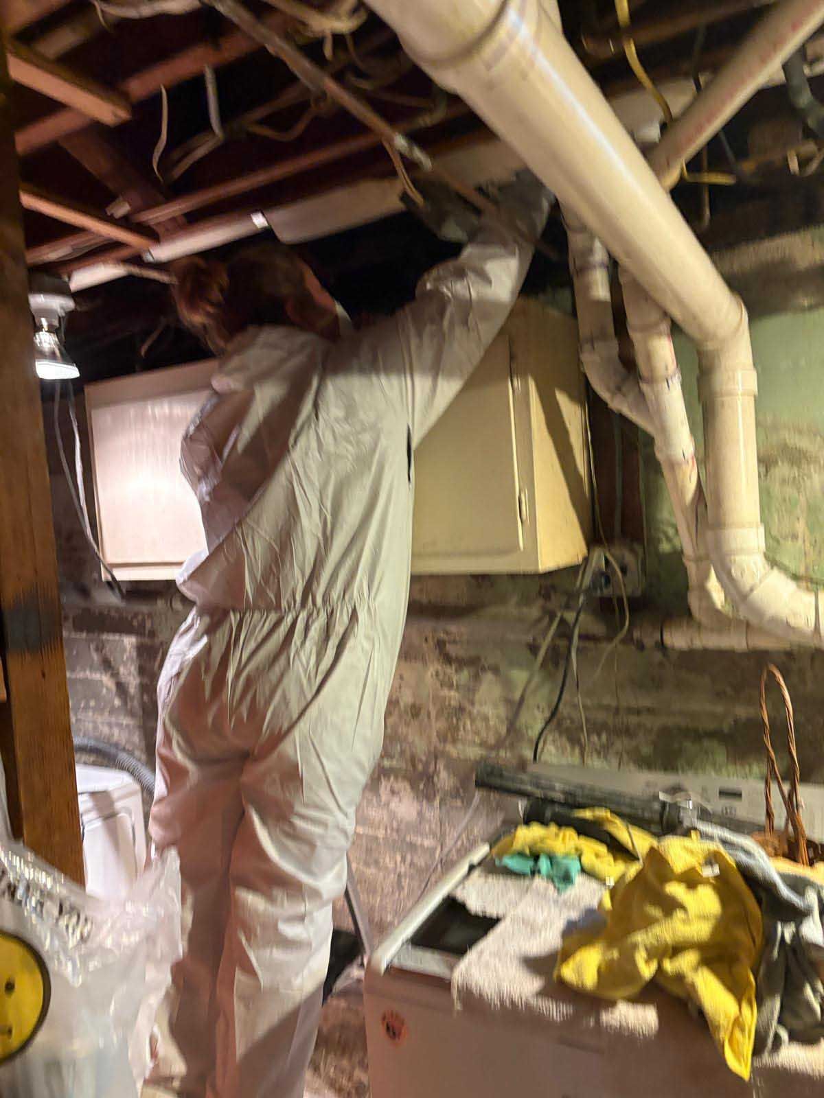 A person in white protective coveralls reaches up to inspect pipes in a dark, unfinished basement.