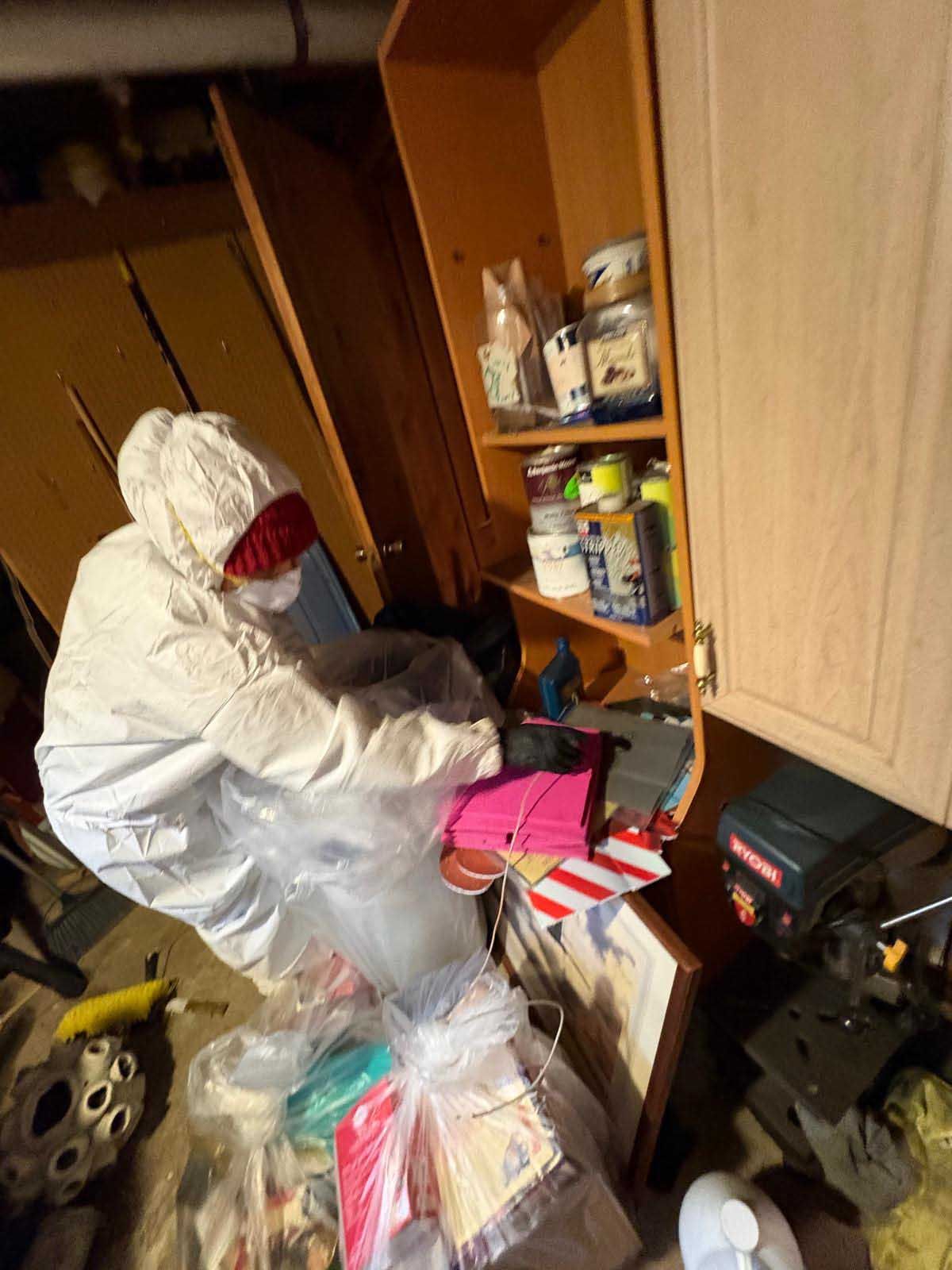 A person in a white hazmat suit and mask clearing items from a shelf into a clear plastic bag in a cluttered basement.
