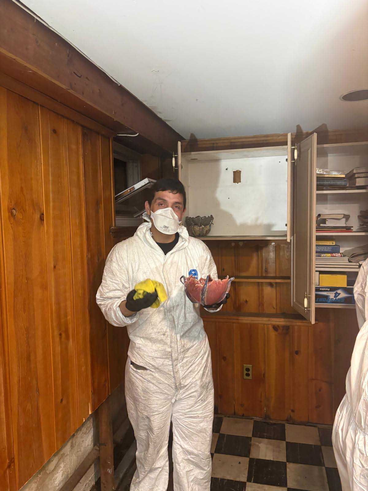 A person in white protective coveralls and a face mask holds a yellow cloth and a small bowl inside a wood-paneled room.