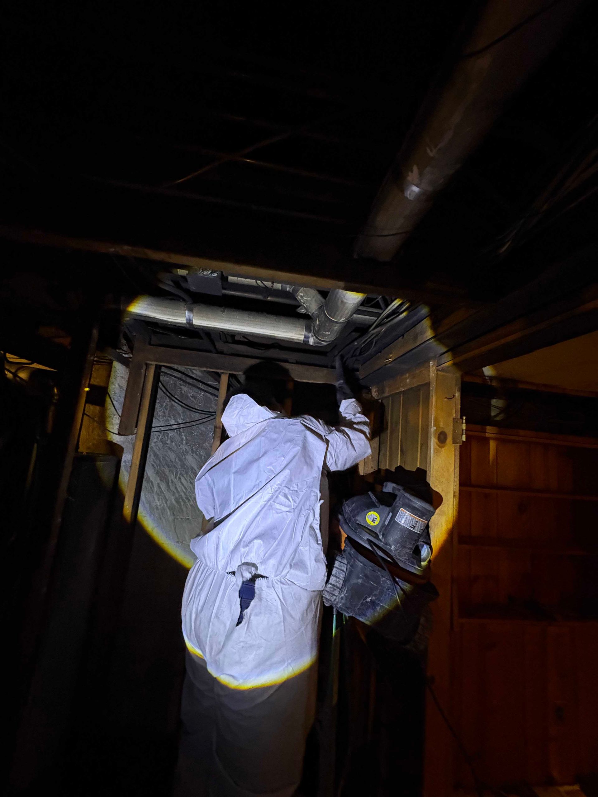A person in white protective coveralls reaching into an open space in a wooden ceiling joist to work on exposed pipes.