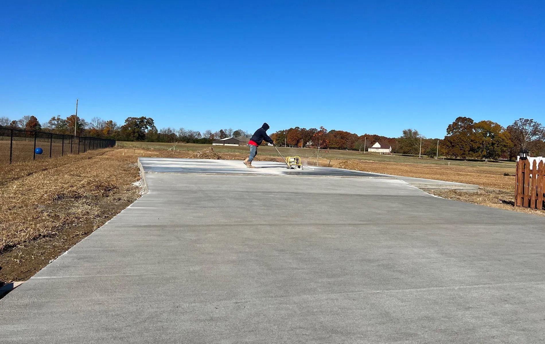 Person on a concrete pad, throwing a ball for a small dog in a field on a sunny day.