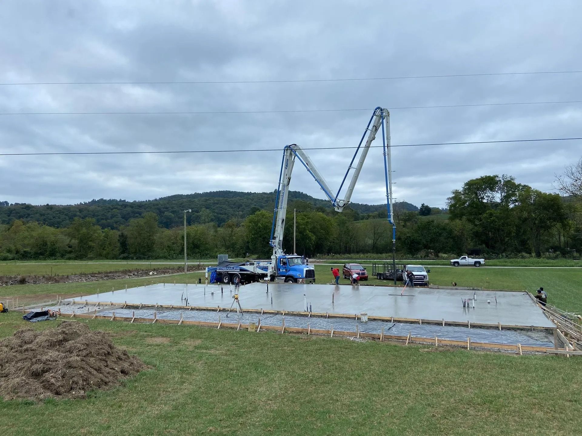 Concrete being poured for a foundation with a pump truck. Cloudy sky, green grass and trees in the background.