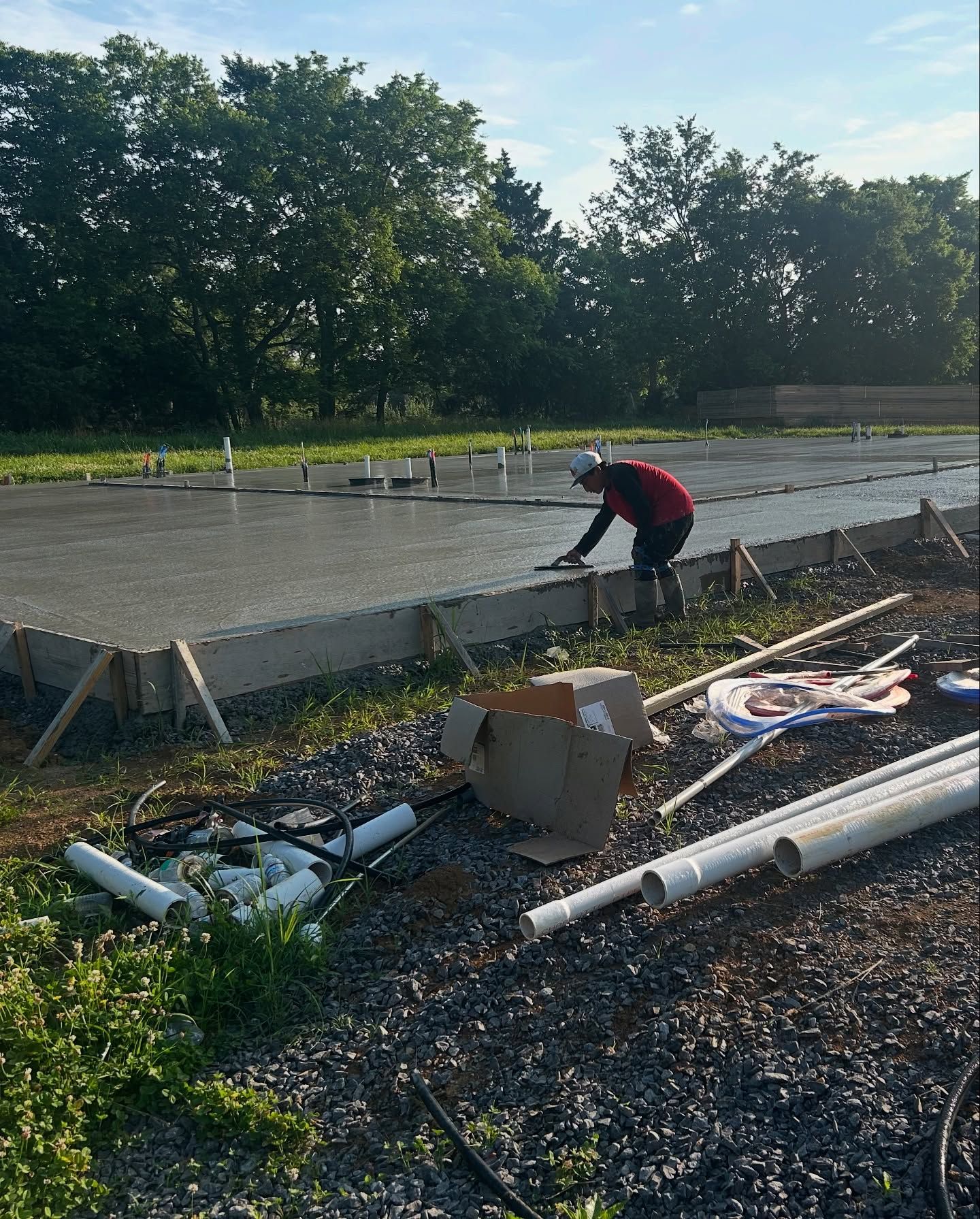 Person smoothing wet concrete on a construction site; lumber frame, pipes, and gravel visible.