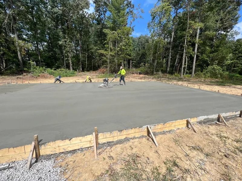 Workers smoothing fresh concrete on a rectangular foundation with a vibratory screed; wooded background.