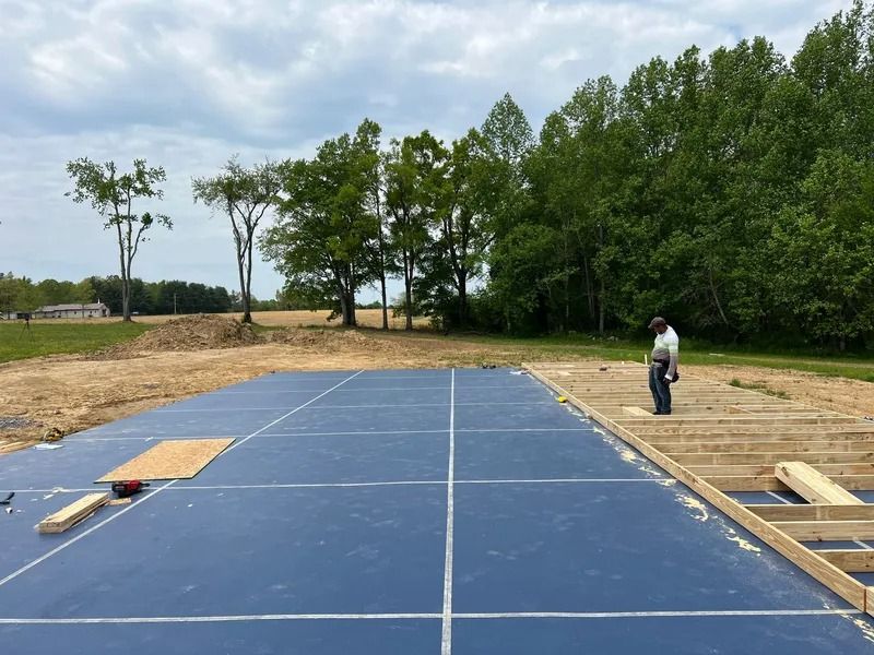 Construction site with a person standing by wooden framing, large blue surface with grid lines, trees in the background.