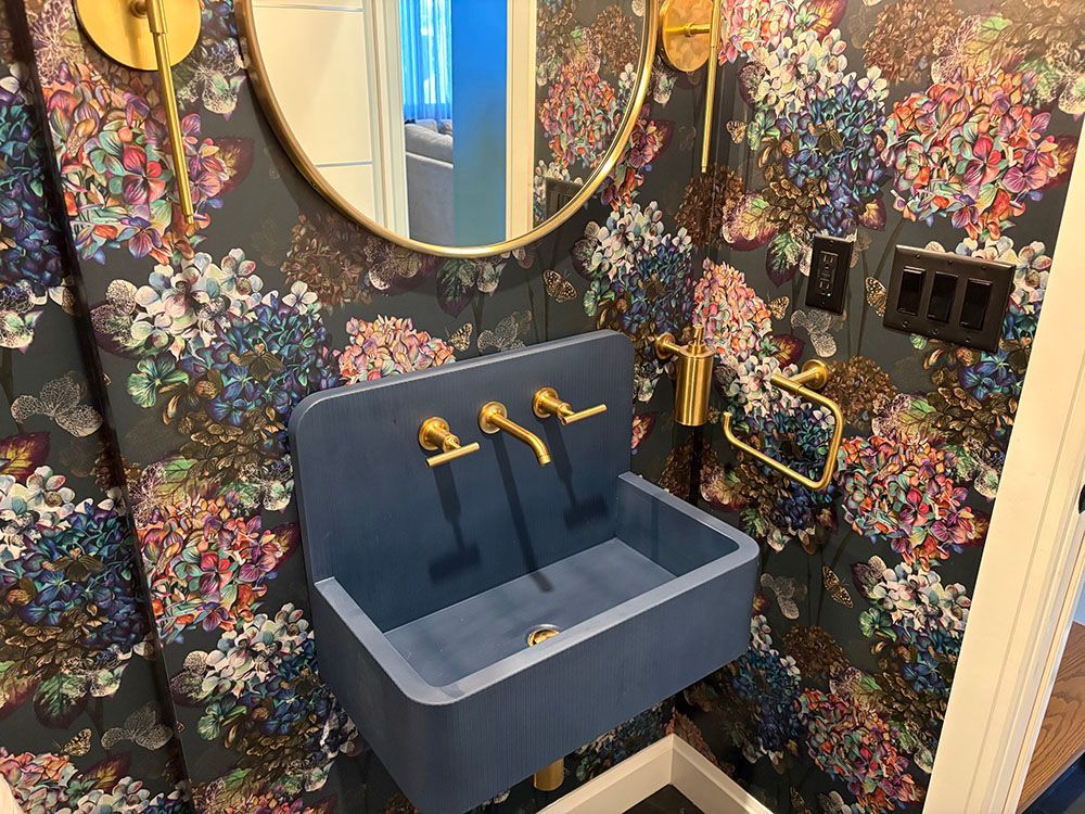 Blue wall-mounted sink in a floral-patterned bathroom with a round mirror and gold faucets.