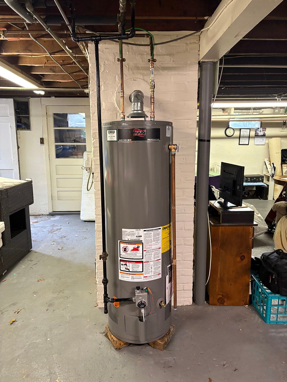 Tall gray water heater in a basement utility area with exposed pipes and ceiling beams.