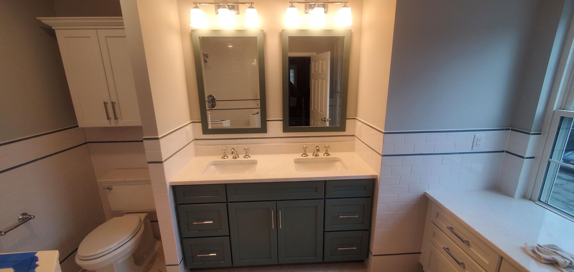 Bathroom with a blue-green vanity, white countertop, mirrors, and lights, next to a toilet and another vanity.
