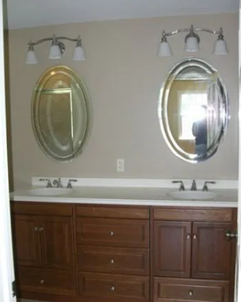 Bathroom with dual sinks, oval mirrors, and dark wood cabinets. Beige walls, chrome lighting fixtures.