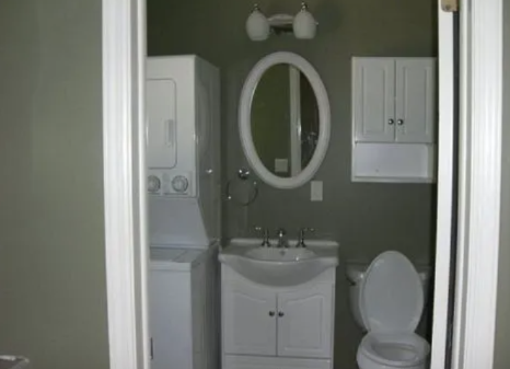 Small bathroom with stacked washer/dryer, oval mirror, sink, toilet, and white cabinets.